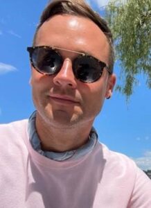 Close-up of Drew wearing a pink sweater, blue bandana around his neck, and sunglasses. 